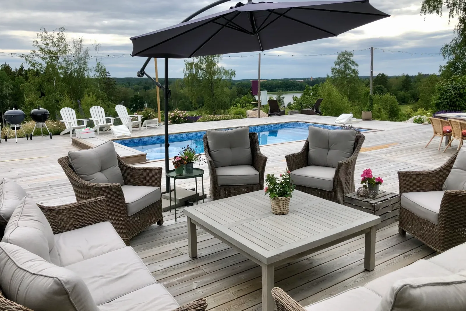 Loungegrupp i rotting under parasoll på trädäcket vid poolen, med grillar och sjöglimt i bakgrunden