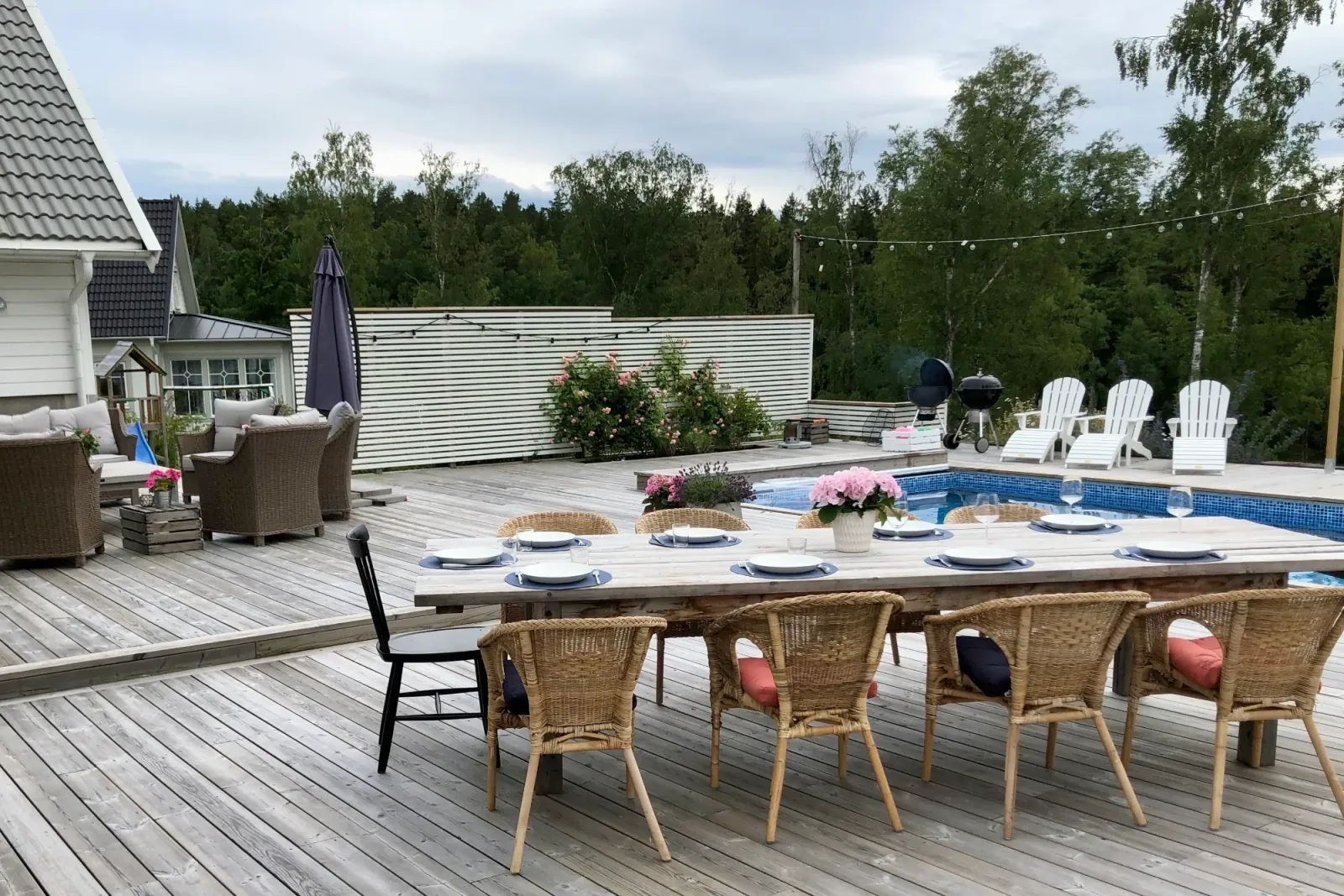 Dukat långbord med rottingstolar på grånat trädäck intill poolen, rosor klättrar på vitt spaljéplank
