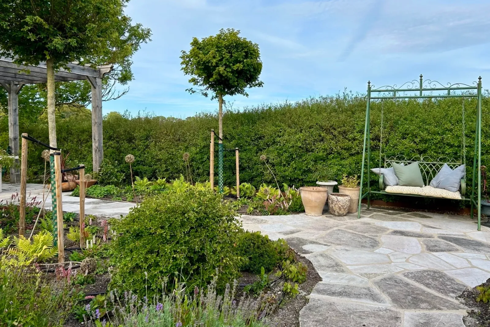 Grön hammock och terrakottakrukor på flislagd kalkstensyta framför hög häck och pergola