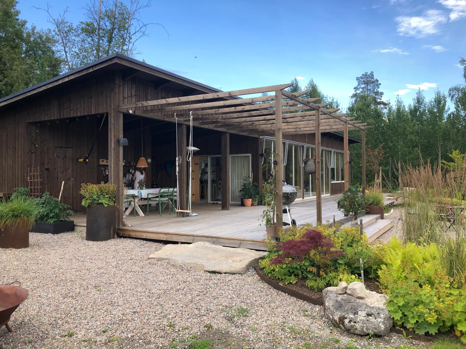 Träpergola sträcker sig längs husets altan med matplats, grill och planteringskärl i rostigt stål
