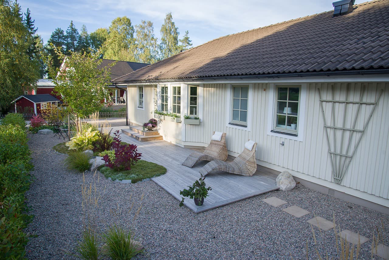 Grusgång längs husfasaden med infällt trädäck, rottingsolstolar och plantering med rödbladig buske
