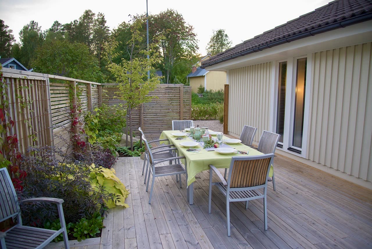 Dukat matbord på trädäck vid husväggen, omgärdat av spjälplank med klätterväxter och lummig rabatt