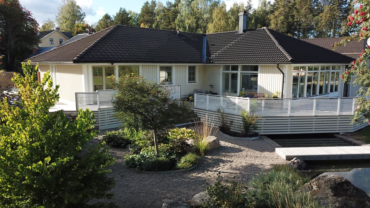 Trädäck med vitmålat spjälräcke som svävar över dammen, träspång och grusade planteringsytor framför huset