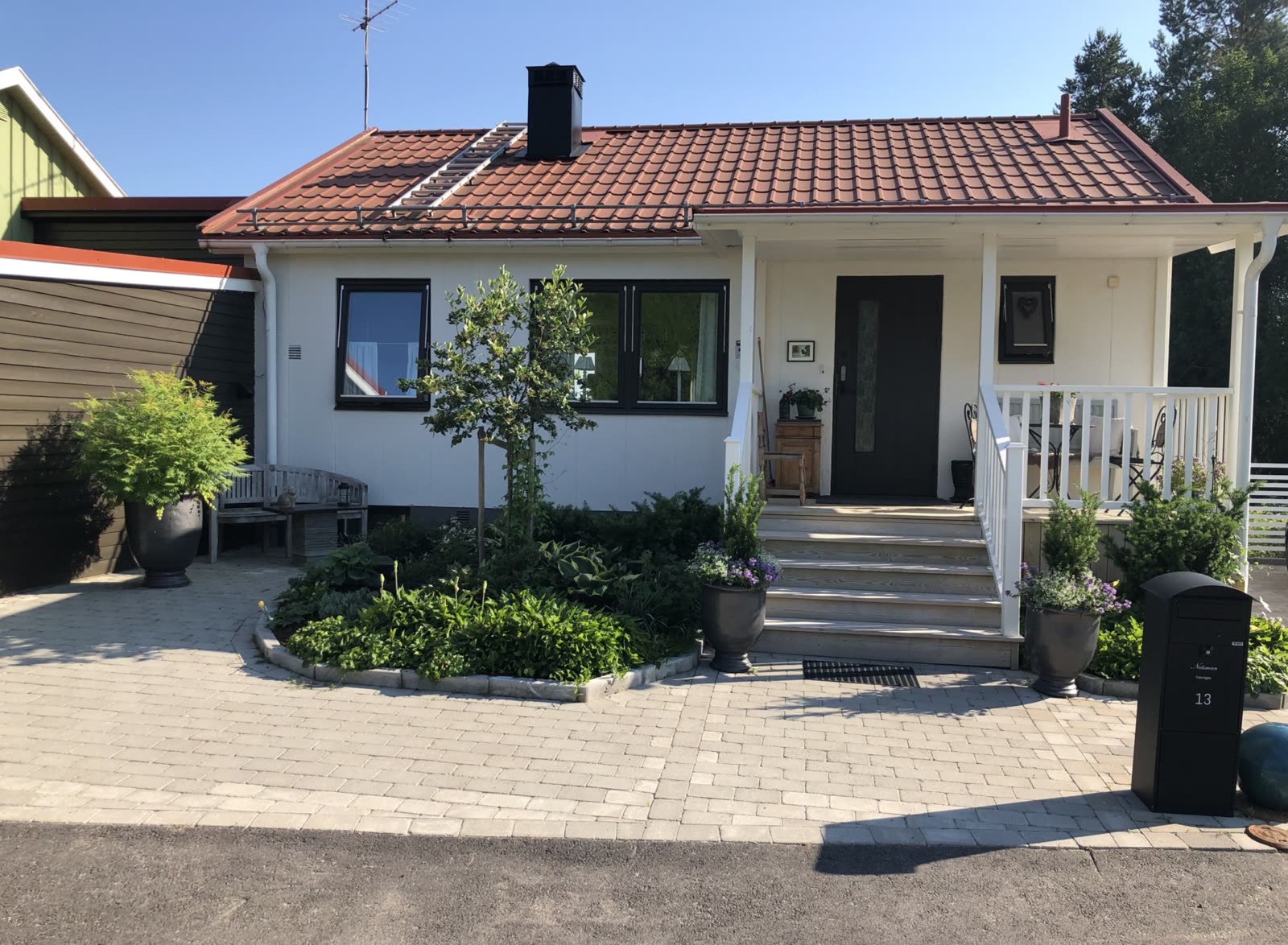 Vitt enplanshus med trätrappa till entrén, plantering med funkia och litet prydnadsträd samt uppfart av betongsten