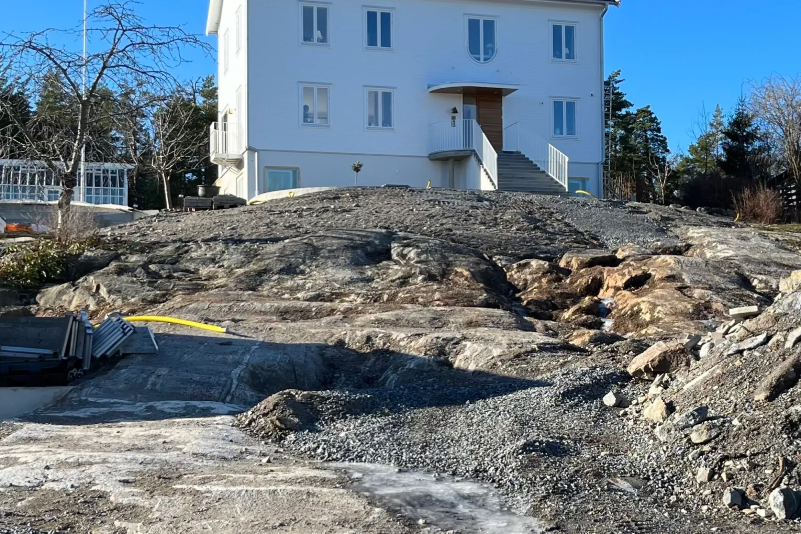 Kal berghäll med grus, slangar och byggmaterial framför nybyggt vitt hus, före anläggning av trädgården