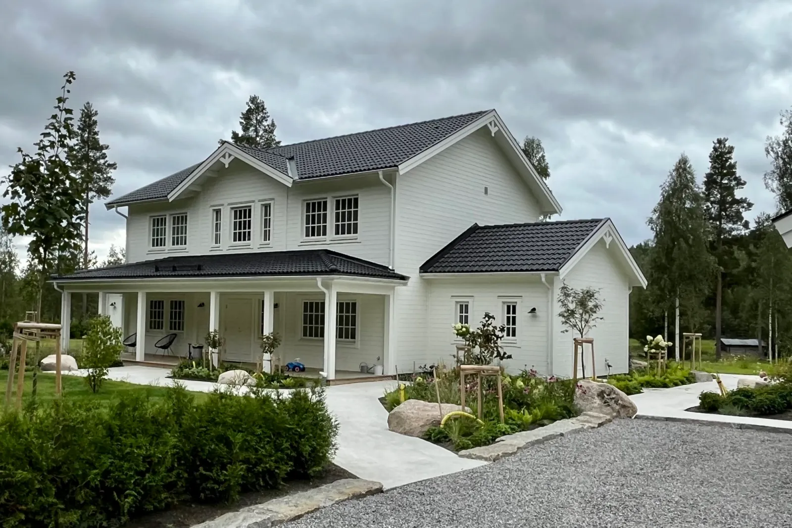 Vit tvåvåningsvilla med svart tegeltak och veranda, omgiven av planteringar med naturstensblock