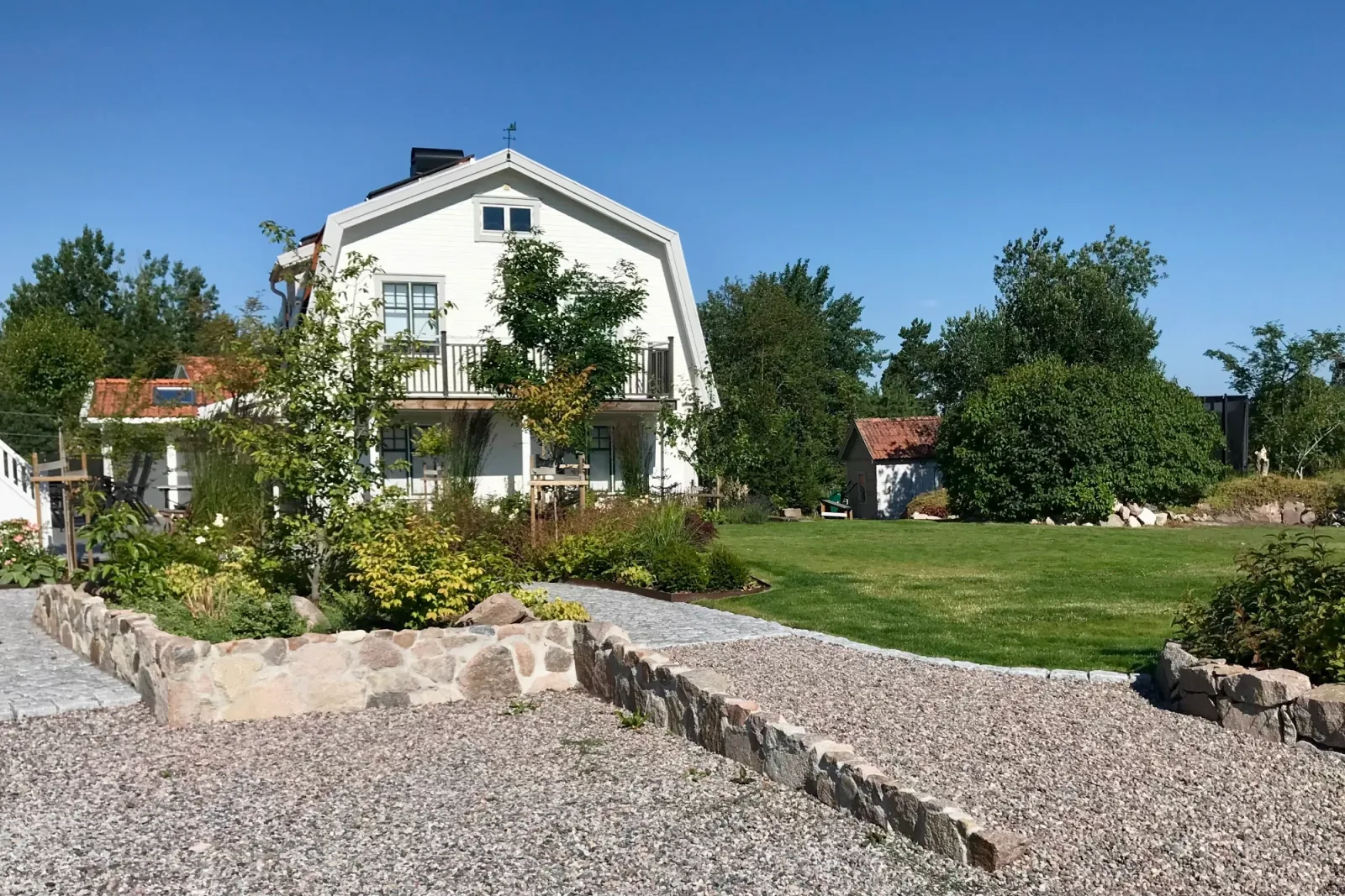 Grusad uppfart avgränsad med låga naturstensmurar, med klippt gräsmatta och vitt tvåvåningshus i fonden